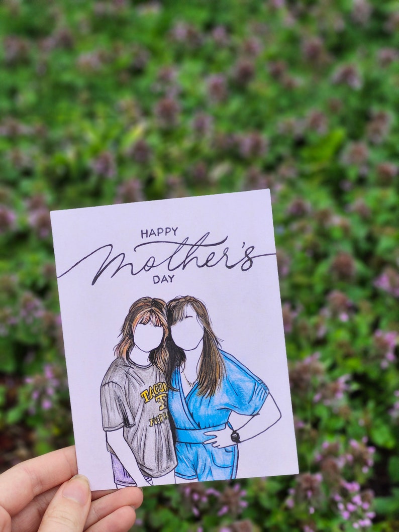 May include: A white card with the text "Happy Mother's Day" in black cursive writing. The card features a hand-drawn illustration of two women standing side-by-side. The woman on the left is wearing a gray t-shirt with the text "TACOMA" in yellow and black. The woman on the right is wearing a blue dress.