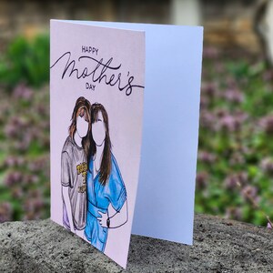 May include: A pink card with the text "Happy Mother's Day" in black script. The card features a hand-drawn illustration of two women, one with brown hair and a blue shirt, and the other with brown hair and a gray shirt.