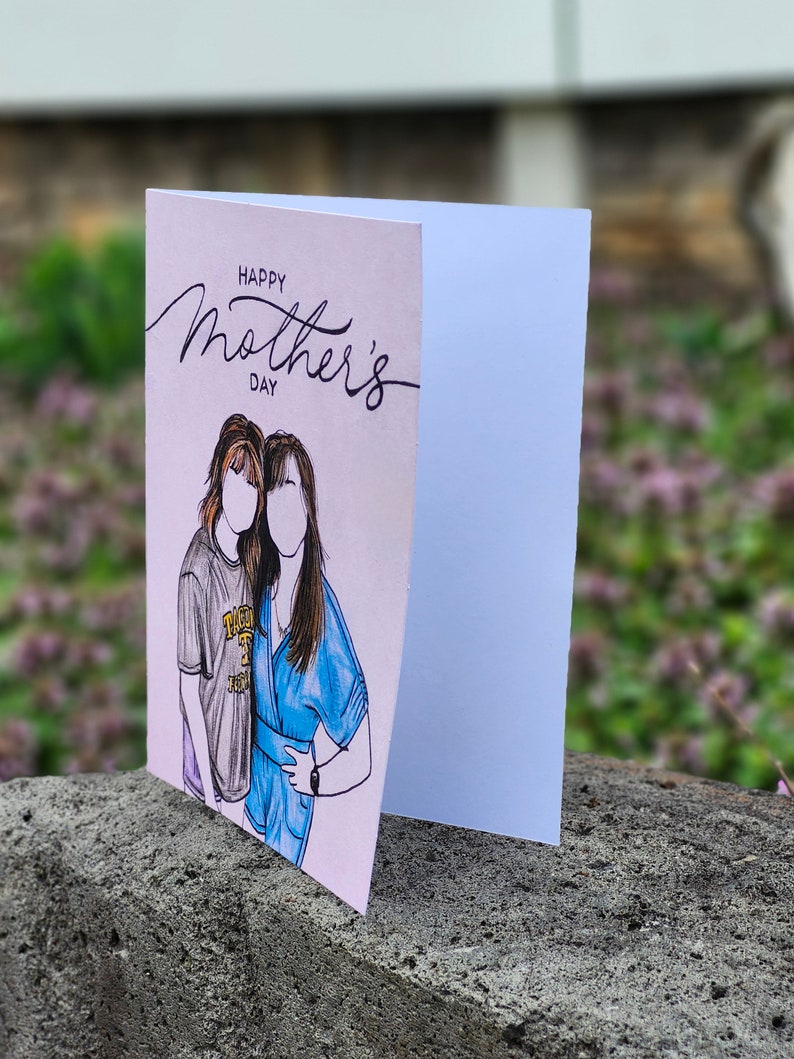 May include: A pink card with the text "Happy Mother's Day" in black script. The card features a hand-drawn illustration of two women, one with brown hair and a blue shirt, and the other with brown hair and a gray shirt.