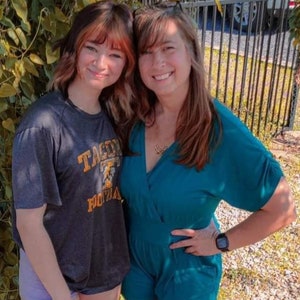 May include: Two women, one wearing a teal blue jumpsuit and the other wearing a gray t-shirt with the text "TACOMA FOOTBALL" printed on it, are standing next to each other. They are smiling and looking at the camera.