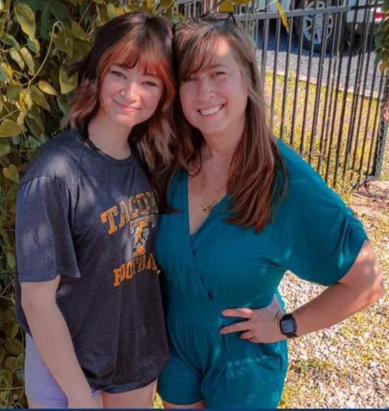 May include: Two women, one wearing a teal blue jumpsuit and the other wearing a gray t-shirt with the text "TACOMA FOOTBALL" printed on it, are standing next to each other. They are smiling and looking at the camera.