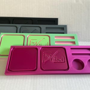 May include: A stack of four rectangular plastic trays in magenta, light green, dark grey, and black. Each tray features two square recessed compartments, two horizontal slots, and two circular cutouts, with the text by Nabell embossed.