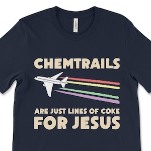 Puede incluir: Una camiseta azul marino con un avión blanco y estelas de arcoíris. El texto "CHEMTRAILS ARE JUST LINES OF COKE FOR JESUS" está impreso en la camiseta.