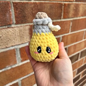 May include: A yellow crocheted potion bottle with a smiling face, black eyes, and pink cheeks. The bottle has a gray and tan stopper and a white curly detail. The plushie is handmade and has a soft, textured appearance.