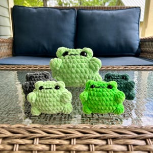 May include: A collection of five handmade crochet frog plushies in various shades of green and gray. The frogs have black eyes and are arranged on a glass table. The plushies are soft and textured, perfect for a nursery or as a gift.