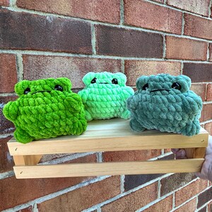 May include: Three plush frog toys made of chunky yarn in shades of bright green, light lime green, and muted sage green. Each frog features a rounded body, small limbs, and two black plastic eyes positioned on the top of the head.