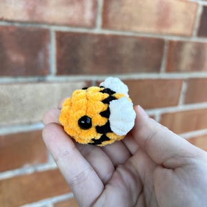 May include: A handmade, plush bee with yellow, black, and white details. The bee has a round body with black stripes, a yellow head, and white wings. The bee's eyes are black. The plush toy is held in a hand.