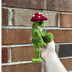 May include: A green crocheted frog plush toy with a textured surface, featuring a dark red mushroom cap with white spots on its head. The frog has large black eyes and a cream-colored chin, crafted from soft yarn with visible stitches throughout.