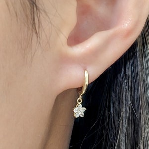 May include: A close-up of a person's ear with a small gold hoop earring with a dangling star-shaped charm set with clear cubic zirconia stones. The text "Size Small" is visible in the lower right corner of the image.