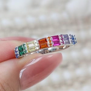 May include: A silver ring featuring a row of colorful rectangular gemstones. The gemstones are arranged in a rainbow pattern, including green, yellow, orange, pink, purple, and blue. The ring is held by a person.