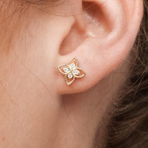May include: A gold-colored stud earring in the shape of a four-petal flower, set with small, clear gemstones at the center of each petal. The earring is worn on an ear, with the person's hair and skin visible.
