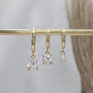 May include: Three gold-colored hoop earrings with dangling, round, clear gemstones of varying sizes. The earrings are displayed on a gold bar against a soft, neutral background. The gemstones sparkle, reflecting light.