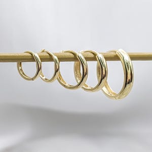 May include: A set of five polished gold hoop earrings arranged in a row from smallest to largest, suspended on a thin horizontal gold bar. Each earring features a smooth, circular design with a hinged closure mechanism visible on the side.