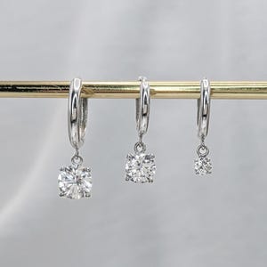 May include: Three silver hoop earrings with dangling round, clear gemstones of varying sizes. The earrings are displayed on a gold-colored bar against a soft, blurred background. The gemstones sparkle, reflecting light.