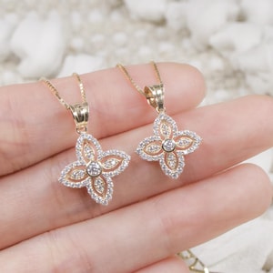 May include: Two gold-toned necklaces with flower-shaped pendants. Each pendant features a central clear stone surrounded by smaller clear stones. The pendants are on delicate gold chains, and the jewelry is displayed on a light-colored surface.