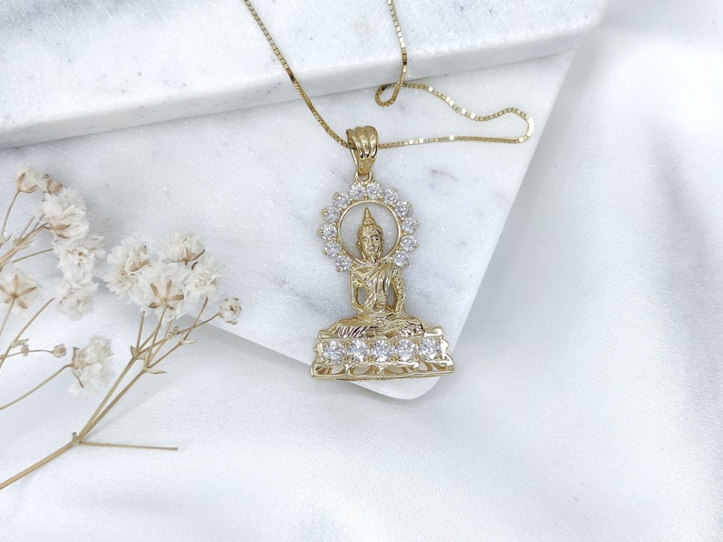 K&ouml;nnte beinhalten: Goldene Halskette mit Anh&auml;nger, der eine detaillierte Buddha-Figur mit einem Heiligenschein aus funkelnden, klaren Steinen zeigt. Der Anh&auml;nger ist an einer zarten goldenen Kette.