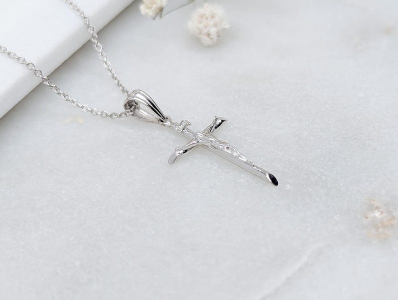 May include: A silver-colored pendant necklace featuring a detailed crucifix design. The pendant is suspended from a delicate chain. The cross depicts a figure and is set against a white background.