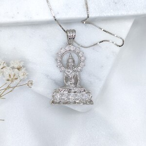 K&ouml;nnte beinhalten: Silberne Halskette mit Anh&auml;nger, der eine detaillierte Darstellung einer sitzenden Buddha-Figur mit einem Heiligenschein aus funkelnden Steinen zeigt. Der Anh&auml;nger ist von einem Kreis aus kleineren Steinen umgeben.