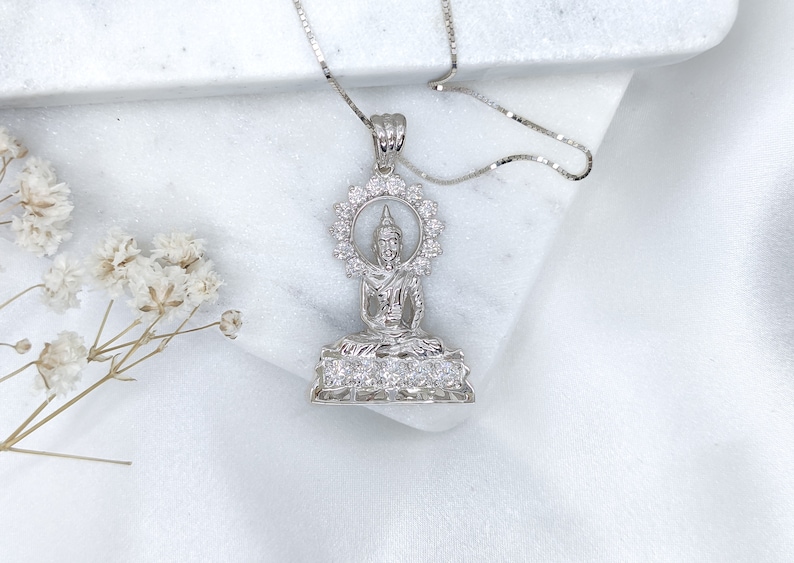 K&ouml;nnte beinhalten: Silberne Halskette mit Anh&auml;nger, der eine detaillierte Darstellung einer sitzenden Buddha-Figur mit einem Heiligenschein aus funkelnden Steinen zeigt. Der Anh&auml;nger ist von einem Kreis aus kleineren Steinen umgeben.