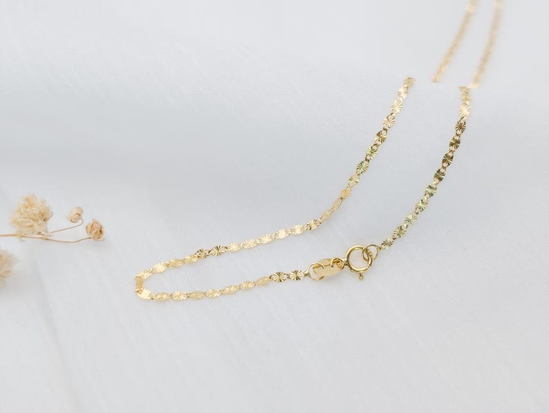 May include: A delicate gold chain necklace with a small clasp. The chain is made of tiny, linked, flat, oval-shaped pieces.