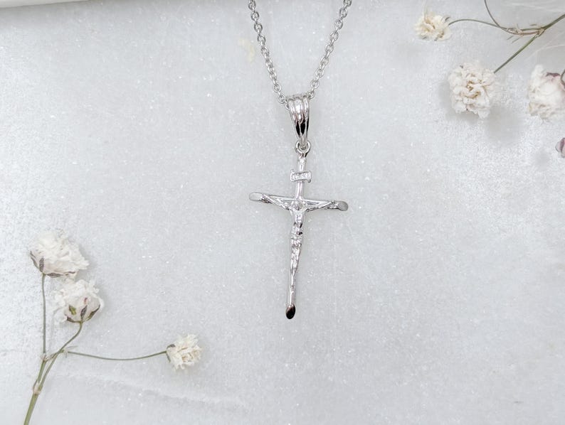 May include: A silver-colored crucifix pendant on a delicate chain. The cross features a detailed depiction of Jesus Christ. The pendant hangs against a white background, with small white flowers in the corners.