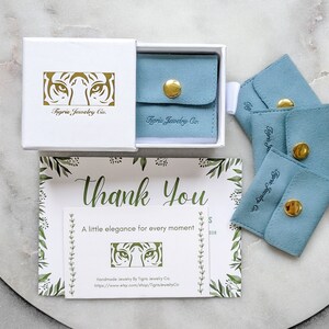 K&ouml;nnte beinhalten: Eine wei&szlig;e Box mit einem goldenen Tigergesichtslogo, auf dem "Tigris Jewelry Co." steht. Die Box ist ge&ouml;ffnet und enth&auml;lt eine kleine blaue Samt-Tasche mit einem goldenen Knopfverschluss. Es gibt vier blaue Samt-Taschen mit goldenen Kn&ouml;pfen und dem aufgedruckten Text "Tigris Jewelry Co.". Eine Dankeskarte mit einem Blumenmuster und dem Text "Danke" und "Ein bisschen Eleganz f&uuml;r jeden Moment" ist ebenfalls sichtbar. Die Karte hat ein kleines goldenes Tigergesichtslogo und den Text "Handgefertigte Schmuckst&uuml;cke von Tigris Jewelry Co. https://www.etsy.com/shop/TigrisJewelryCo."