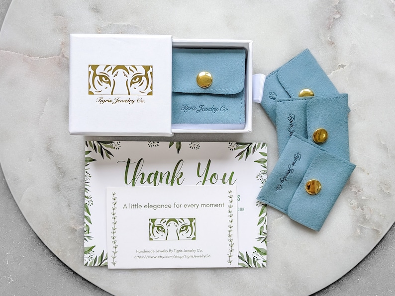 K&ouml;nnte beinhalten: Eine wei&szlig;e Box mit einem goldenen Tigergesichtslogo, auf dem "Tigris Jewelry Co." steht. Die Box ist ge&ouml;ffnet und enth&auml;lt eine kleine blaue Samt-Tasche mit einem goldenen Knopfverschluss. Es gibt vier blaue Samt-Taschen mit goldenen Kn&ouml;pfen und dem aufgedruckten Text "Tigris Jewelry Co.". Eine Dankeskarte mit einem Blumenmuster und dem Text "Danke" und "Ein bisschen Eleganz f&uuml;r jeden Moment" ist ebenfalls sichtbar. Die Karte hat ein kleines goldenes Tigergesichtslogo und den Text "Handgefertigte Schmuckst&uuml;cke von Tigris Jewelry Co. https://www.etsy.com/shop/TigrisJewelryCo."