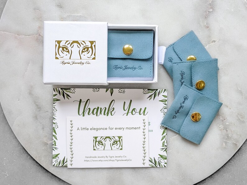 Puede incluir: Una caja blanca con un logotipo de cara de tigre dorado que dice "Tigris Jewelry Co." La caja est&aacute; abierta y contiene una peque&ntilde;a bolsa de terciopelo azul con un cierre de bot&oacute;n dorado. Hay cuatro bolsas de terciopelo azul con botones dorados y el texto "Tigris Jewelry Co." impreso en ellas. Una tarjeta de agradecimiento con un dise&ntilde;o floral y el texto "Gracias" y "Un poco de elegancia para cada momento" tambi&eacute;n es visible. La tarjeta tiene un peque&ntilde;o logotipo de cara de tigre dorado y el texto "Joyer&iacute;a hecha a mano por Tigris Jewelry Co. https://www.etsy.com/shop/TigrisJewelryCo."