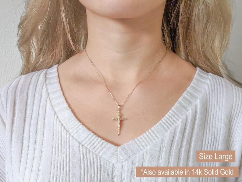 May include: A gold-colored cross pendant necklace. The cross features a detailed depiction of Jesus. The necklace is displayed on a person wearing a white ribbed sweater. Text on the image reads "Size Large" and "Also available in 14K Solid Gold."