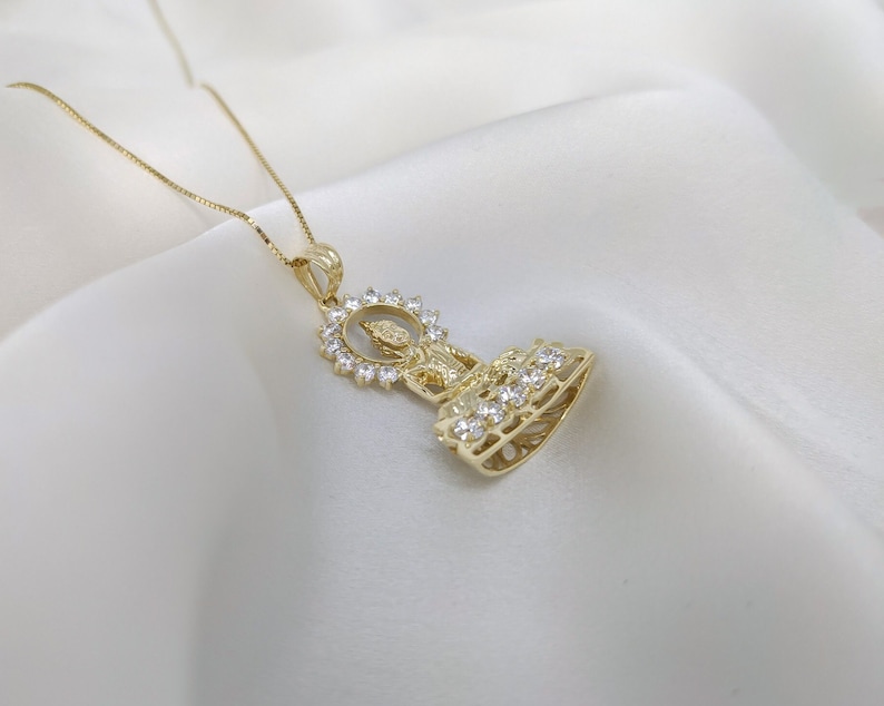 K&ouml;nnte beinhalten: Eine goldene Halskette mit einem Anh&auml;nger, der eine detaillierte Figur eines sitzenden Buddha zeigt, umgeben von funkelnden, klaren Steinen.