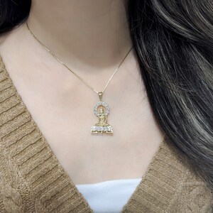 K&ouml;nnte beinhalten: Eine goldene Kettenkette mit einem Anh&auml;nger, der eine sitzende Buddha-Figur zeigt, die von einem Heiligenschein aus klaren Steinen umgeben ist.
