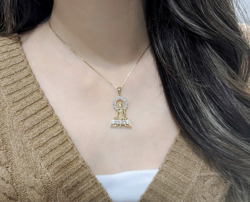 K&ouml;nnte beinhalten: Eine goldene Kettenkette mit einem Anh&auml;nger, der eine sitzende Buddha-Figur zeigt, die von einem Heiligenschein aus klaren Steinen umgeben ist.