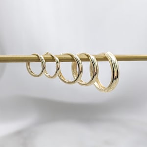 May include: A set of five gold hoop earrings arranged in a row from smallest to largest, hanging on a thin horizontal gold rod. Each earring features a smooth, polished finish and a circular design with a hinged closure mechanism for secure wear.