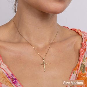 May include: A gold-colored crucifix pendant necklace. The pendant features a detailed depiction of Jesus on the cross. The necklace is shown on a person. Text on the image says "Size Medium" and "Also available in 14k Solid Gold."