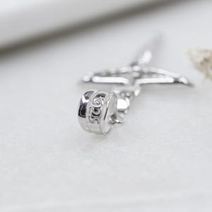 May include: Close-up of a silver-colored cross pendant with a detailed design. The pendant has a textured surface and is set against a soft, blurred background. The word "DIOR" is visible on the pendant.
