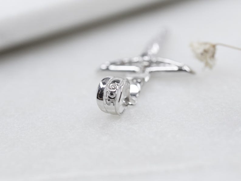 May include: Close-up of a silver-colored cross pendant with a detailed design. The pendant has a textured surface and is set against a soft, blurred background. The word "DIOR" is visible on the pendant.
