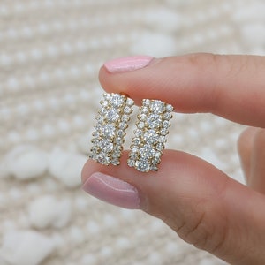 May include: A pair of gold earrings with a row of small, clear cubic zirconia stones. The earrings are in the shape of a rectangle and are held in a hand.