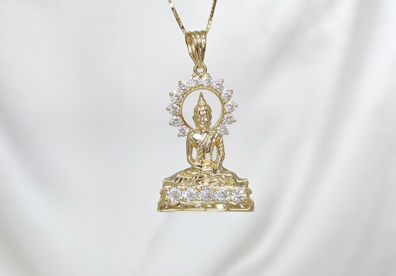 K&ouml;nnte beinhalten: Goldene Halskette mit Anh&auml;nger, der eine detaillierte Buddha-Figur mit einem Heiligenschein aus klaren Zirkonia-Steinen zeigt. Der Anh&auml;nger befindet sich auf einem wei&szlig;en Hintergrund.