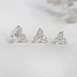 May include: Three pairs of diamond stud earrings in varying sizes. Each earring features a cluster of three round-cut diamonds, set in a silver or white gold metal. The earrings are displayed on a white surface, showcasing their sparkle and design.