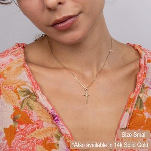 May include: A delicate gold-colored cross pendant necklace. The cross features a detailed depiction of Jesus Christ. The necklace is shown on a person wearing a floral patterned top. The text "Size Small" is visible.