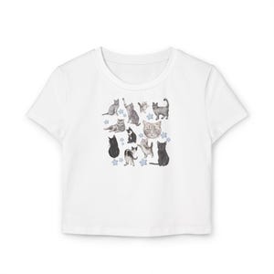 May include: White cropped t-shirt with a print of various cats in different poses. The cats are in shades of gray and black, and there are small blue stars scattered around them.