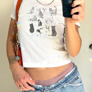 May include: A white cropped t-shirt with a graphic print of various cats and stars. The shirt is being worn by a person with a gold chain necklace and a brown leather bag.