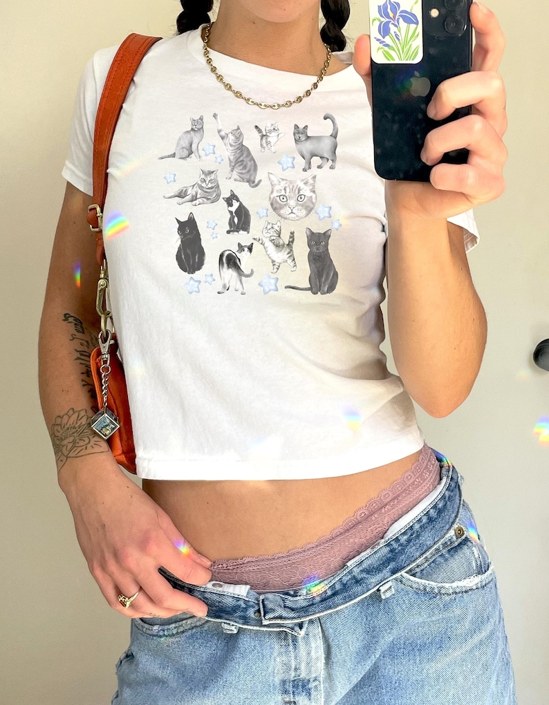 May include: A white cropped t-shirt with a graphic print of various cats and stars. The shirt is being worn by a person with a gold chain necklace and a brown leather bag.