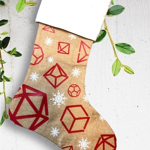 May include: A Christmas stocking with a burlap texture, featuring red geometric shapes and white snowflakes. The top of the stocking is white, and it hangs from a beige loop. The design includes various polyhedral dice shapes.
