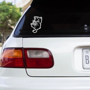 Può includere: Un'auto bianca con un adesivo di un gatto nero sul lunotto posteriore. Il gatto tiene un cuore nelle sue zampe.