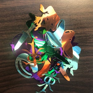 May include: A collection of colorful, metallic, laser-cut shapes, including a frog, a sword, and a leaf. The shapes are layered on top of each other and have a shiny, iridescent finish.