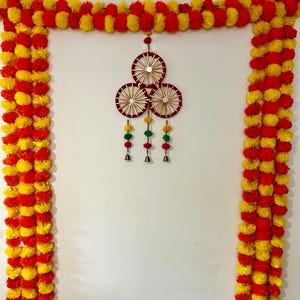 May include: A decorative door hanging featuring long garlands of alternating yellow and red pom-poms. Centered is a wall ornament composed of three circular frames with gold spokes and mirrors, accented by hanging pom-poms and small brass bells.
