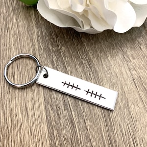 May include: Silver keyring with a rectangular tag engraved with a series of stitches. The keyring is on a wooden surface.