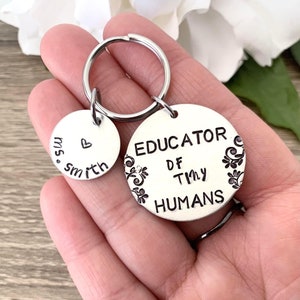 May include: A silver keyring with two round tags. The larger tag says "EDUCATOR OF TINY HUMANS" with a decorative design. The smaller tag says "ms. smith" with a heart symbol.
