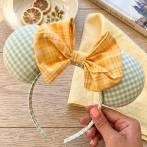 May include: A handmade headband with mouse ears. The ears are green and white gingham, and the bow is yellow and orange plaid. The headband is green and white gingham. A book titled "Magical Memories - Castles & Dreams" is in the background.
